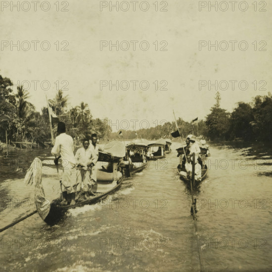 Some boats on the Sungai Martapura river, with both Europeans and original inhabitants of Borneo, stereograph, inscription: 'Terrain exploration Marta-, poera river', Sungai Martapura, 1900 - 1940, photographer, Borneo, paper, pasteboard, baryta paper, gelatin silver print, developing gelatin silver print, height 90 mm x width 179 mm, river, A sepia-toned scene showing narrow wooden boats moving along a river with people standing and sitting in them, some holding long poles and small dark flags, while canopies cover portions of several boats; the riverbanks are lined with dense tropical trees and tall palms, and gentle ripples trail behind the boats across the water surface.