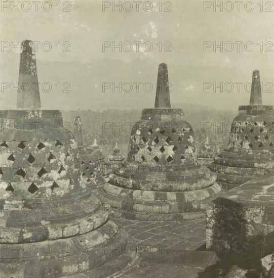 Stupas on Borobudur, Java, Boroboedoer, stereograph, Borobudur, 1900 - 1940, photographer, Central Java, paper, pasteboard, baryta paper, gelatin silver print, developing gelatin silver print, height 90 mm x width 185 mm, places and objects of worship, Hinduism, Buddhism, Jainism, Stone stupas with conical pinnacles and perforated bell-shaped bodies fill the foreground, their surfaces mottled with lichen-like patches and weathering; layered circular bases and stone walkways with visible joints and wear surround them, and a hazy distant landscape of treetops and faint mountain silhouettes recedes under a pale, lightly speckled sky.