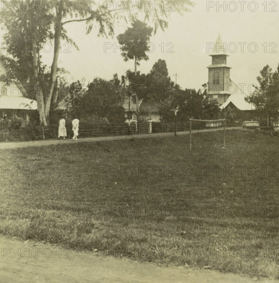 Path to a church with two people in Tondano, Indonesia, stereograph, inscription: 'Minahasja (Manado), Tondano', Tondano, 1900 - 1940, photographer, Celebes, Noord-Celebes, paper, pasteboard, baryta paper, gelatin silver print, developing gelatin silver print, height 87 mm x width 175 mm, village street, Roman Catholic Church, North Celebes, A wide grassy foreground leads to a dirt path with two figures in light clothing walking near a fence, trees with tall trunks and broad canopies frame the scene, and a multi-tiered tower with a pointed roof rises above surrounding buildings and foliage.