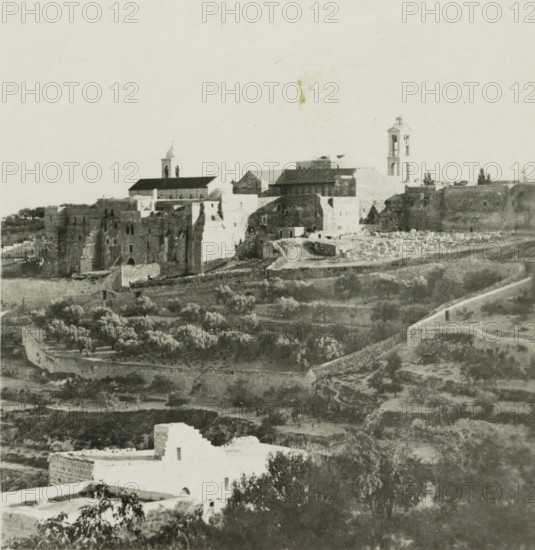 Bethlehem, View of Bethlehem, stereograph, address: 'Parklaan 49, HAARLEM, (Holland)', inscription: 'These stereo-photos are being offered for sale and sold wholesale and retail by the ........., ', number: '166.', monogram, inscription: 'Déposée.', photographer: Richard Sochor, publisher: De Wereld-Tourist, Haarlem, 1903, Palestine, paper, baryta paper, gelatin silver print, gelatin silver developing print, height 88 mm x width 179 mm, city-view in general, 'veduta', A wide panoramic view shows an old stone cityscape with a cluster of large masonry buildings and fortified walls on a hill, two prominent bell towers rising above the roofline, pitched roofs and flat terraces visible among the structures, and a road or path winding along the base of the walls. In the foreground there are terraced fields and groves with rows of trees separated by low stone walls, scattered smaller buildings with whitewashed facades, and irregular patches of bare earth and vegetation. The overall image is m...