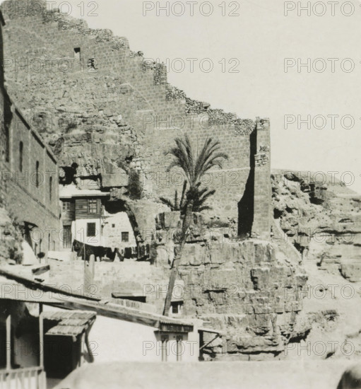 Vallée du Cédron. Le couvent Mar Saba, Mar Saba monastery in the Kidron Valley, Israel, stereograph, inscription: 'Déposée.', address: 'Parklaan 49, HAARLEM, (Holland)', number: '189.', inscription: 'These stereo photographs are marketed and sold wholesale and retail by the ........., ', monogram, photographer: Richard Sochor, Mar Saba, publisher: De Wereld-Tourist, Haarlem, 1903, Jerusalem, Palestine, Kidron-dal, paper, baryta paper, gelatin silver print, developing gelatin silver print, height 90 mm x width 178 mm, abbey, monastery, convent, Roman Catholic church, A stone fortress wall with stepped crenellations rises against a pale sky above a rocky cliff face, with rough stone blocks and rubble visible along the wall and cliff; near the base of the wall there is a tall narrow vertical shadowed opening and a slender palm tree with feathery fronds stands in front of the rock; adjacent to the cliff are small buildings with stone and plaster facades, wooden shutters, a balcony, and a l...
