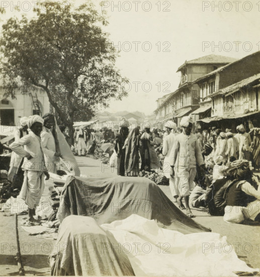 Bazaar in India, stereograph, inscription: 'Bazaar at', India, 1900 - 1920, photographer, paper, pasteboard, baryta paper, gelatin silver print, developing gelatin silver print, height 88 mm x width 176 mm, market, A busy outdoor market scene with many people wearing light-colored traditional garments and headwraps moving among stalls and goods spread on the ground, a row of covered bundles and draped fabrics running down the center foreground, vendors and customers standing, walking, crouching, and carrying items on their heads, a large leafy tree on the left side, multi-story buildings with sloped roofs and awnings lining the right side, and a crowded street filled with activity extending into the distance.