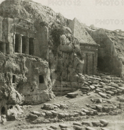 Jerusalem Sépulcres St. Jacques et St. Zachari, Rock Tombs of Benei Hezir and St. Zacharia, Kidron Valley, stereograph, number: '159.', inscription: 'Déposée.', monogram, address: 'Parklaan 49, HAARLEM, (Holland)', inscription: 'These stereo photographs are marketed and sold wholesale and retail by the ........., ', photographer: Richard Sochor, Kidron-dal, publisher: De Wereld-Tourist, Haarlem, 1903, Jerusalem, paper, baryta paper, gelatin silver print, developing gelatin silver print, height 88 mm x width 179 mm, grave, tomb, Tomb of Benei Hezir, Tomb of Zechariah, A grayscale photograph of a rocky cliff face with several rectangular carved openings and recessed columns set into the rock, adjacent to a stepped stone pathway and scattered blocks; the rock surfaces show weathered textures and erosion, with a triangular rock formation above one carved recess and uneven rocky ledges and small cavities visible across the scene.