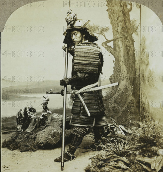 Ancient warrior costume of the Japanese. Japan, Japanese soldier in traditional dress, stereograph, copyright notice: 'Copyright 1902 by CH Graves.', number: '30', number: '59', number: '190.', photographer: T. Enami (possibly), Japan, publisher: Universal Photo Art Company, Philadelphia, 1900, North America, paper, pasteboard, baryta paper, gelatin silver print, developing gelatin silver print, height 89 mm x width 178 mm, the soldier, the soldier's life, A sepia-toned portrait of a person wearing layered armor and a decorated helmet, standing with a long pole held vertically and a sheathed sword at their side; the armor shows woven and plated textures with straps and lacing visible, and the helmet has an ornate crest and flared neck guard. The person looks toward the camera with a neutral facial expression, one hand gripping the pole and the other resting near the sword hilt. The scene includes a painted backdrop with a tree trunk and distant landscape, and the foreground shows rocks...