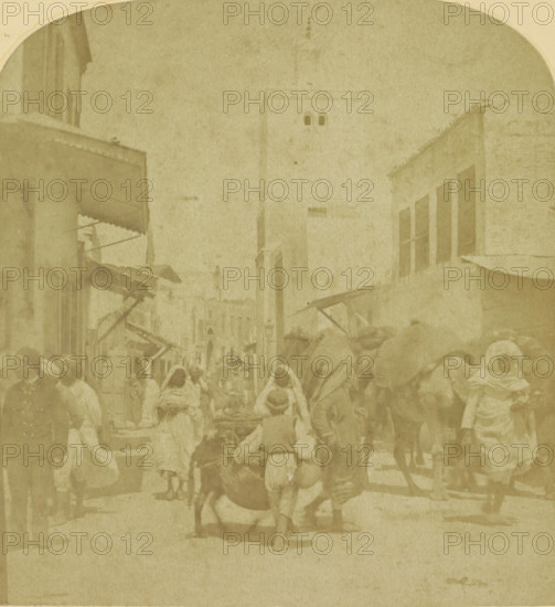 Street scene in an Arab city, possibly in Morocco, stereograph, inscription: 'C', number: '25175', inscription: 'SPECIAL ADDRESS FOR GOOD STEREOSCOPES, AND BEAUTIFUL STEREOSCOPIC PHOTOS.', photographer: August Frederik Willem Vogt, Morocco (possibly), 1897 - 1922, North Africa, pasteboard, paper, albumen print, height 88 mm x width 179 mm, street, A faded sepia street scene showing a narrow urban thoroughfare flanked by multi-story buildings with shuttered windows and awnings, a tall tower rising in the background, and many pedestrians wearing loose draped garments and head coverings moving along the street; some figures lead or ride animals, while others carry bundles or push carts, and market stalls with angled canopies extend from the building fronts.
