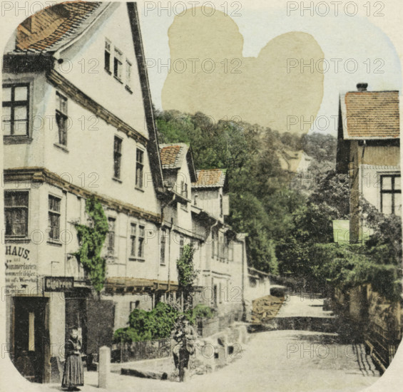 Harz. Das Gasthaus auf dem Hexentanzplatz, Hotel on the Hexentanzplatz, Harz, stereograph, number: 'No. 12.', inscription: 'Der Hexentanzplatz, Treppensteig von 1100 Stufen. (W.Riehl)', inscription: 'Hof-Photograph. Potsdam.', photographer: Hermann Selle, Hexentanzplatz, 1866 - 1870, pasteboard, paper, albumen print, albumendruk, height 83 mm x width 170 mm, hotel, hostelry, inn, mountain-top, peak, A narrow street scene with rows of multi-story buildings lining one side, their light-colored facades interspersed with dark window frames and tiled roof sections, a taller building with a steep roof visible toward the right, and leafy green trees and foliage rising behind and between the structures; a person stands near the foreground wearing a striped skirt and dark top, another figure is further down the street near a low stone wall, and the street surface leads to a sunlit open area framed by shadows and balustrades.