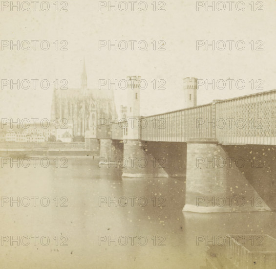 Pont fixe et Cathédrale, Dombrücke over the Rhine and Cologne Cathedral, Germany, Cologne (Prusse), Allemagne, stereograph, inscription: 'Déposé.', number: 'N. 543', inscription: '(Déposé)', photographer: Hippolyte Jouvin, 1864, North Rhine-Westphalia, pasteboard, paper, albumen print, height 86 mm x width 177 mm, church (exterior), bridge, Cologne Cathedral, Dombrücke, A faded sepia photograph shows a large iron bridge spanning a river, supported by stone piers rising from the water, with patterned metal railings and towers with crenellated tops at the near end; behind the bridge a Gothic cathedral with a tall pointed spire and detailed façade is visible above a row of buildings along the riverbank, and the water in the foreground reflects the bridge piers and has scattered spots and discolorations across the image.