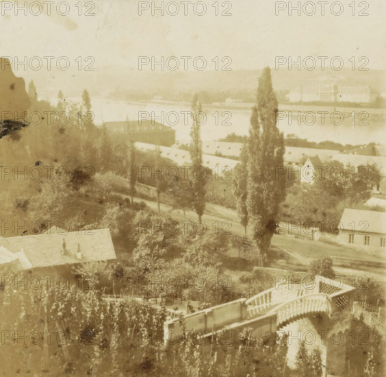 Confluent of the Moselle and the Rhine at Coblentz, Meeting of the Moselle and the Rhine near Koblenz, Germany, Boards of the Rhine, stereograph, inscription: '6', number: 'No 76', Koblenz, 1861 - 1870, photographer, Rhineland-Palatinate, pasteboard, paper, albumen print, height 86 mm x width 172 mm, river, city-view, and landscape with man-made constructions, Rhine, Moselle, A faded sepia view showing a riverside scene with tall narrow trees rising above rooftops and garden foliage, a long low building with a sloped roof near the water, a winding path or road passing among trees, a pale body of water stretching across the middle distance, distant low hills beyond the water, and portions of decorative wooden railings and terraces in the foreground.