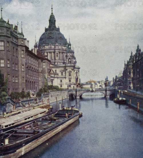 View of the Spree and the Stadschloss in Berlin, Spree am Klg. Schloss., La Sprée devant le Château royal., The Spree at the Royal Palace., La Sprea devanti al Castello Reale., El Spree cerca del Palacio Real, Berlin, photomechanical print, stereograph, inscription: 'Warenzeichen, No. 142123', inscription: 'Wir sthen auf der Kurfürstenbrüchke und, ', inscription: 'Chromoplast-Bild, No. 56', inscription: 'Serie 7 Berlin I No. 55-60', inscription: 'DRGM, No. 437756', inscription: '2.', cliché maker: Farbenphotographischen Gesellschaft, Stuttgart, 1911 - 1919, photographer, Baden-Württemberg, paper, pasteboard, three-color car type, height 90 mm x width 181 mm, bridge in city across river, canal, palace, A riverside scene with a large domed building dominating the left center, flanked by multi-story stone buildings with rows of windows and decorative roof spires, a stone bridge crossing the river beneath the dome, small boats moored along the near quay with wooden hulls and open decks, a ...