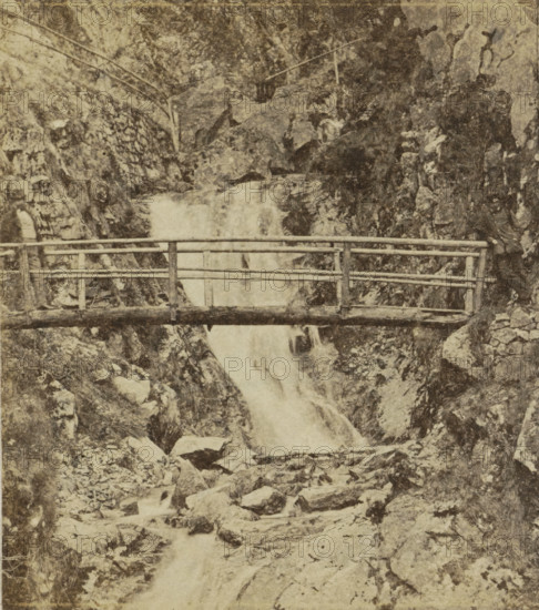 All Saints' Waterfall near Oppenau, Germany, All Saints' Day, stereograph, photographer: Numa Blanc, Oppenau, 1857 - 1867, Baden-Württemberg, pasteboard, paper, albumen print, height 86 mm x width 174 mm, A weathered wooden footbridge spans a rocky gorge with a waterfall flowing beneath it; the bridge has horizontal rails and vertical supports and appears aged. Jagged rock faces and boulders surround the gorge on both sides, with rough textured surfaces and patches of lighter and darker stone. Water cascades down over the rocks, creating a foamy, lighter-toned stream as it descends. Sparse vegetation and tufts of grass are visible among the rocks and along the edges of the gorge. Two human figures stand at opposite ends of the bridge, partially visible and wearing dark clothing, with one figure leaning against the railing. The overall image has a high-contrast, grainy, sepia-toned appearance and shows surface marks and spots.