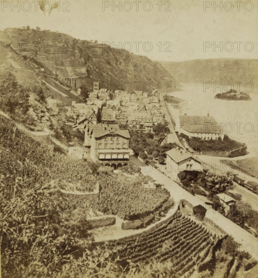 View of Bacharach, Germany, Bacharach, Der Rhein und seine Umgebung, The Rhine and his environ, stereograph, photographer: Anselm Schmitz, 1877, Rhineland-Palatinate, pasteboard, paper, albumen print, height 88 mm x width 178 mm, village, dale, valley, A sepia-toned scene showing a hillside town beside a wide river with steep terraced slopes rising behind the cluster of buildings; rows of pitched-roof houses and larger multi-story structures line a narrow road that runs parallel to the river, and a tower-like building stands among the rooftops; an island of rock sits in the river near the middle right, while terraced vineyards or cultivated plots cover the foreground slope with winding paths and stone walls; trees and shrubs dot the hillside and village, and a few larger buildings with arched openings are visible close to the riverbank.