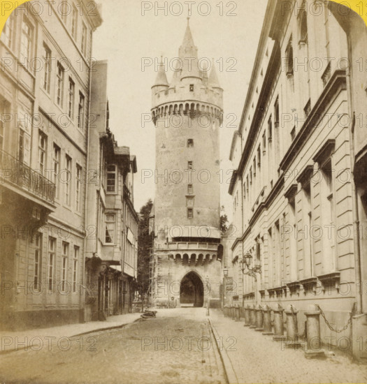 Tour d'Eseinheim à Francfort, Eschenheimer Turm in Frankfurt am Main, Bords du Rhin, stereograph, inscription: ', 4', number: 'No 211.', inscription: '11062, eg, ', Frankfurt am Main, 1858 - 1863, photographer, Hessen, pasteboard, paper, albumen print, height 86 mm x width 172 mm, street, city-gate, Eschenheimer Turm, A narrow street lined with tall multi-story stone buildings on both sides leads to a large cylindrical stone tower with battlements and conical turrets at the top; the tower has a large arched opening at its base and a balcony-like projection with a row of corbels beneath it, and there is scaffolding against one side of the tower; the street surface is cobbled with a raised sidewalk and a row of short stone posts linked by chains along one side, and ornate wall-mounted lanterns are attached to the building facades.