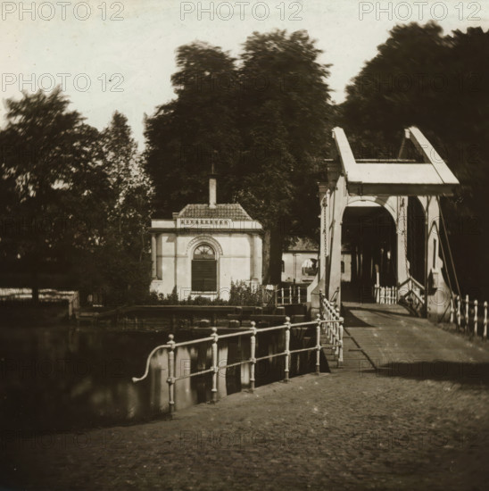 View of 's Gravelandsche poort in Weesp, stereoscopic photograph, address: 'NZ Voorburgwal, bij het Spui.', photographer: Pieter Oosterhuis, Weesp, 1856, Pieter Oosterhuis, photographer, inorganic material, glass, slide, height 85 mm x width 173 mm, bridge, A waterfront scene showing a small classical building with a tiled roof and a narrow chimney, an arched window with shutters on its facade, dense dark trees rising behind it, and a white wooden drawbridge with curved support beams and hanging chains spanning a canal; in the foreground a metal railing runs along the canal edge above water, a paved walkway leads toward the bridge, and reflections appear in the water beside the quay.