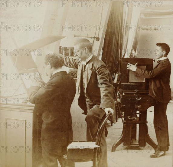 Photographers at work in a studio, stereograph, number: '12', Netherlands, 1882 - 1920, photographer, paper, pasteboard, daylight gelatin silver print, height 88 mm x width 179 mm, photographer at work, A sepia photograph showing three men in a room with tall windows and heavy curtains; one man seated at a window holding a magnifying glass to a flat surface, another man standing beside him leaning forward with one hand resting on the seated man's shoulder and the other on the back of a chair, and a younger man standing near a large wooden camera or box apparatus with his hands on its side. The men wear dark suits with high collars and cravats, and the room contains wooden furniture including a turned chair and a stand supporting the camera, as well as cylindrical containers on a shelf and visible window frames and moulding. The lighting is bright from the left, casting soft highlights and shadows across the scene, and the image has an aged, scratched texture.