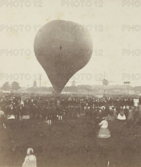 Ascent of Nadar's hot air balloon 'le Géant' on September 14, 1865, Giant balloon 'le Géant' by Nadar is launched in Amsterdam, September 14, stereograph, inscription: 'The Govert Flinckstraat has been cleared on this site., Amsterdam.', retailer's mark: 'BOOKSTORE, JAN D. BROUWER., KALVERSTRAAT E No 37, AMSTERDAM.', number: '10', photographer: Pieter Oosterhuis, Amsterdam, publisher: Jan Dirk Brouwer, 1865-09-14, pasteboard, paper, albumen print, height 85 mm x width 176 mm, balloon (aviation), A large teardrop-shaped balloon fills the center of the scene, its surface smooth and slightly mottled; a dense crowd of people stands and sits across the middle ground, with some figures closer to the foreground visible from the back and wearing long skirts or coats; in the distant background there are silhouettes of windmills and a few tall structures, and a long low fence or row of tents stretches horizontally; the overall image has a sepia tone and an aged photographic texture with faint sp...