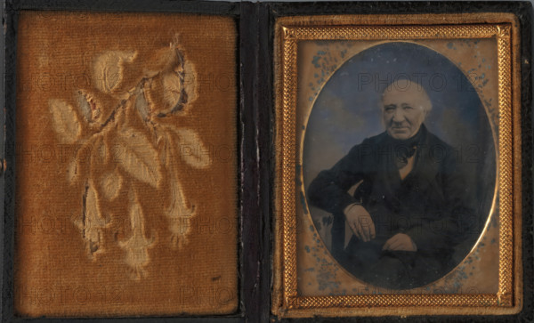 Portrait of an unknown man, photograph, c. 1855 - c. 1870, photographer, plant material, textile materials, biological material, inorganic material, glass, metal, velvet (fabric weave), wood (plant material), leather, ambrotype, in wooden case covered with leather with velvet lining, height 55 mm x width 44 mm, height 74 mm x width 62 mm x thickness 15 mm x width 126 mm, historical persons, sitting figure, A rectangular case opened to show two panels, the left panel lined with worn golden-brown velvet embossed with a faded floral design, the right panel containing an oval photograph encased in a decorative gold-colored frame with textured border and small ornamental corners; the photograph shows an older man seated, wearing a dark jacket and scarf, with one arm resting on an armrest and the other hand visible near his lap, his hair light and receding and his expression neutral, while age spots and speckles are visible on the photographic surface and the surrounding mat.