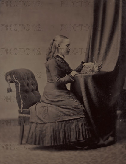 Portrait of an unknown young woman sitting at a table with a cat, photograph, 1855 - 1900, photographer, metal, inorganic material, iron (metal), ferrotyping, height 100 mm x width 68 mm, historical persons - BB - woman, adolescent, young woman, maiden, cat, sitting figure - AA - female human figure, A seated young woman in a long dress seen in profile with her hair pulled back and her hands resting on a draped table where a striped cat lies, a padded chair with tassels beside her, and a heavy curtain hanging behind the table.