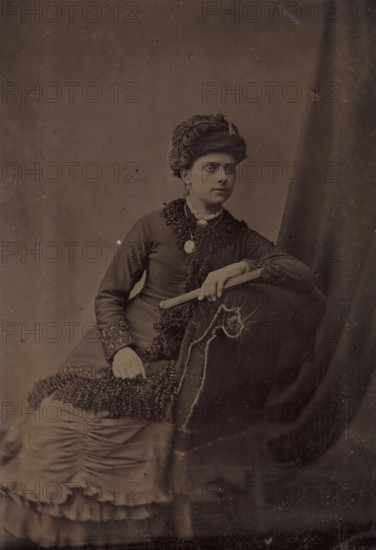 Portrait of an unknown woman with a hat, photograph, 1855 - 1900, photographer, metal, inorganic material, iron (metal), ferrotyping, height 104 mm x width 69 mm, historical persons - BB - woman, head-gear: cap, A seated woman wearing a dark dress with textured trim and a high collar is shown against a plain backdrop, she has an ornate hat with layered fabric and a decorative element, her hair is styled beneath it and she wears earrings and a brooch at her throat, one arm rests on an upholstered armrest with visible seam details while the other hand holds a slender folded object, the lower part of her dress shows gathered and layered fabric with ruffled edges, the image has a subdued monochrome tone and scattered surface blemishes.