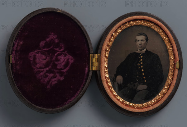 Portrait of an unknown man, possibly a soldier, photograph, union case, 1861, photographer, textile materials, organic material, inorganic material, metal, glass, velvet (fabric weave), plastic, ferrotyping, height 68 mm x width 56 mm x thickness 17 mm, anonymous historical person depicted, An open oval case with a hinged center showing a mirrored pair of interiors, the left side lined with deep purple velvet featuring a raised floral pattern and a dark rimmed edge, and the right side containing an oval photographic plate framed by a peach-colored padded border and a decorative gold inner trim, the photograph showing a seated man with short hair wearing a dark buttoned coat and a light-colored bow at the collar, his hand resting on his knee and the image surface showing small specks and slight wear.