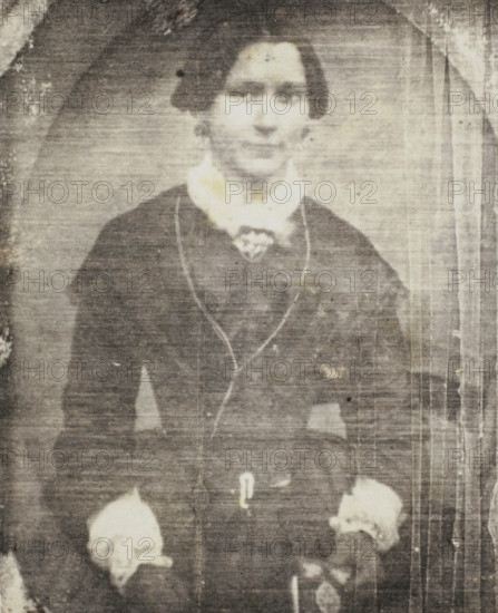 Photo reproduction of a daguerreotype of a portrait of an unknown woman, photograph, reproduction, France, after photo by anonymous, 1840 - 1860, photographer, paper, salted paper print, height 72 mm x width 59 mm, adult woman, historical persons not known by name - BB - woman, A faded black-and-white portrait of a person from the waist up wearing a dark dress or coat with a light-colored high collar and decorative trim, with hands resting at the sides holding light-colored floral objects, a faint oval backdrop around the head, and vertical streaks and spots from age or wear across the image.