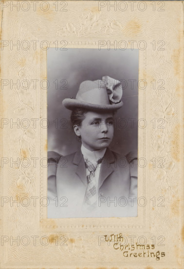 Christmas greeting with portrait of an unknown woman with a hat, photograph, Christmas card, Portrait of an unknown woman with a hat with the text 'With Christmas Greetings'., inscription: 'With, Christmas, Greetings', England, 1890 - 1910, photographer, paper, pasteboard, baryta paper, gelatin silver print, pressing, developing gelatin silver print, height 95 mm x width 67 mm, height 60 mm x width 33 mm, adult woman, anonymous historical person depicted, A sepia-toned portrait of a woman set within a textured ornate frame, showing her from the chest up wearing a tailored jacket with puffed sleeves, a high-collared blouse, and a patterned necktie; she wears a hat with a rounded crown and a decorative folded ribbon on one side, and her hair is styled back from her face; her expression is neutral as she looks slightly away from the camera; the frame around the photograph has embossed floral motifs and wavy textured patterns and shows some areas of discoloration.