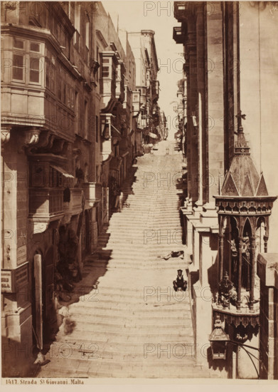 Strada S. Giovanni in Valetta, Malta, Strada St. Giovanni. Malta, photograph, inscription: '(+), Sommer', number: '1417', photographer: attributed to Giorgio Sommer, Valetta, 1857 - 1914, Attributed to, pasteboard, paper, albumen print, albumendruk, height 354 mm x width 270 mm, sloping street, street with steps or stairs, A long, steep stone staircase runs upward between tall buildings on both sides, with narrow balconies and enclosed wooden bay windows projecting from the facades. The left side shows ornate stone corbels and recessed doorways while the right side features a decorative stone canopy with pointed arches and a small sculptural figure beneath it. Shadows fall across the steps, where a few people are seated or standing, and narrow shopfronts and awnings line the lower portions of the buildings. The street appears sunlit toward the top of the stairs and darker in shadow near the bottom.