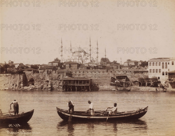 View of the Sultan Ahmet Mosque in Istanbul, Mosquée du Sultan-Ahmed, photograph, number: 'N, 429.', photographer: Sébah & Joaillier, Istanbul, 1888 - 1900, paper, albumen print, height 208 mm x width 269 mm, temple, shrine, Islam, Mohammedanism, Sultan Ahmet Mosque, A waterfront scene shows small wooden boats on calm water with three men in one boat engaging with nets and oars while another man stands in a nearby boat. Behind them a stone seawall and tiered buildings stretch along the shore, including a stilted wooden structure extending over the water. Above the shoreline a large domed building with multiple slender spires rises behind rows of low houses and trees under a pale sky.