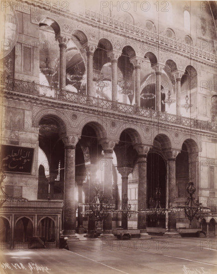 Interior of the Hagia Sophia in Istanbul, Ste Sophie, photograph, number: 'N. 478.', photographer: Sébah & Joaillier, Istanbul, 1888 - 1900, paper, albumen print, height 269 mm x width 210 mm, temple, shrine, Islam, Mohammedanism, Hagia Sophia, A large interior space framed by rows of tall stone columns with ornate capitals supporting a series of arched openings and an upper gallery with decorative balustrades, intricate carved patterns along the arches and spandrels, multiple hanging chandeliers of varying designs suspended at different heights, high vaulted ceilings with visible archways and painted or textured surfaces, a polished floor with visible seams, low wooden partitions and archways at one side, a few benches and raised platforms scattered near the columns, and soft light filtering through the openings creating highlights and shadows across the architectural details.