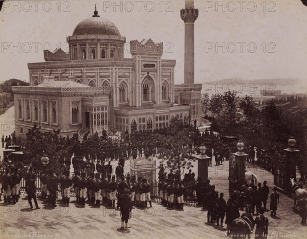 Ceremony of Islam, Crowd during the reception of Sultan Abdülhamit II for the Salat al-djuma (Friday afternoon prayer) at the Yildiz Hamidiye Mosque in Istanbul, photograph, number: '689', photographer: Abdullah Frères, Istanbul, 1876 - 1909, paper, albumen print, height 200 mm x width 259 mm, temple, shrine, Islam, Mohammedanism, crowd, mob, Yildiz Hamidiye Mosque, A large ornate building with a domed roof and a tall slender tower stands on a raised terrace overlooking a body of water, its façades featuring arched windows, decorative cornices, and latticework panels. In front of the building a crowd of uniformed men is gathered along a gated entrance and the terrace steps, some standing in formation while others cluster near carriages and horses. Trees and shrubs frame parts of the scene, and lamp posts with spherical glass tops appear near the entrance. Boats and distant shoreline structures are visible across the water behind the building, and the overall image has a sepia tone.