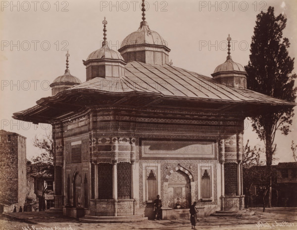 Fountain of Sultan Ahmed III in Istanbul, Fontaine Ahmed, photograph, number: '163.', photographer: Sébah & Joaillier, Istanbul, 1888 - 1900, paper, albumen print, albumendruk, height 207 mm x width 267 mm, utilitarian, public pump or water fountain, Topkapi Palace, Fountain of Sultan Ahmed III, A stone building with ornate carved panels and patterned tile work has a wide overhanging roof and multiple small domes topped with finials; the roof shows segmented metal panels and decorative edges, and the facade features arched doorways, recessed windows with lattice screens, and detailed relief borders; two figures stand near the entrance, one closer to the doorway and another a short distance away holding a stick or pole, and a tall tree rises beside the building while another smaller tree and adjacent structures are visible at the sides.