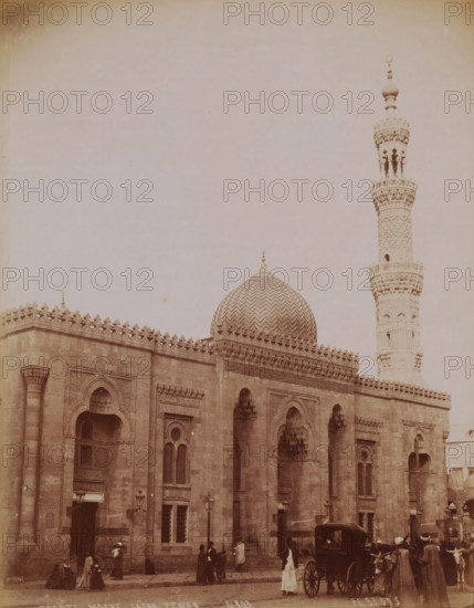 Al-Sayeda Zainab Mosque in Cairo, Crante Moske Saïda Zenab, photograph, number: 'N 303', photographer: Peridis, Cairo (city), 1870 - 1900, Ottoman Empire, North Africa, paper, albumen print, height 281 mm x width 220 mm, temple, shrine, Islam, Mohammedanism, Al-Sayeda Zainab Mosque, A sepia-toned photograph showing a large ornate stone building with a patterned domed roof and a tall carved minaret rising beside it, the building facade featuring arched windows and doorways with decorative stonework and crenellated parapet detailing, street-level scene with groups of people wearing period clothing gathered and walking near the entrance, a horse-drawn carriage parked in front, and a few street lamps along the sidewalk.
