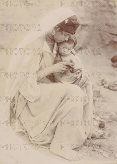 Portrait of an unknown woman with child, photograph, Middle East, 1860 - 1900, photographer, paper, albumen print, height 290 mm x width 214 mm, anonymous historical person portrayed, mother and baby or young child, A seated woman wearing a draped light-colored garment and a sheer veil over her head holds a small child on her lap; the woman has a wrapped headscarf with decorative elements and layered necklaces, and she looks down toward the child while holding the child's hand; the child is partially wrapped in cloth, faces forward with a neutral expression, and has a small head covering; both figures show visible hands and the woman's bare foot rests on the ground amid a rough, sandy surface with scattered stones.