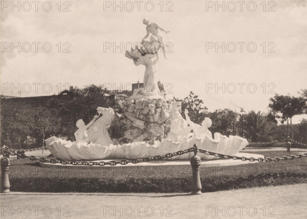 Nereids fountain in Buenos Aires, Fuente Lola Mora, Paseo de Julio, photomechanical print, Buenos Aires, cliché maker: anonymous, after sculpture by Lola Mora, 1904 - 1905, Lola Mora, photographer, after sculpture by, pasteboard, paper, collotype, photoprint, height 167 mm x width 227 mm, height 259 mm x width 332 mm, ornamental fountain, A light-colored sculpted fountain sits on a circular base with a low stone rim and a ring of heavy chains supported by short posts around it; the fountain features a central rocky column topped by a large shell holding a crouching male figure and a standing female figure beneath, with additional sculpted figures and horse heads emerging from shell-like basins at the lower level; the scene is set outdoors with trimmed grass in the foreground, trees and shrubbery behind the fountain, and a partly cloudy sky filling the upper area of the image.