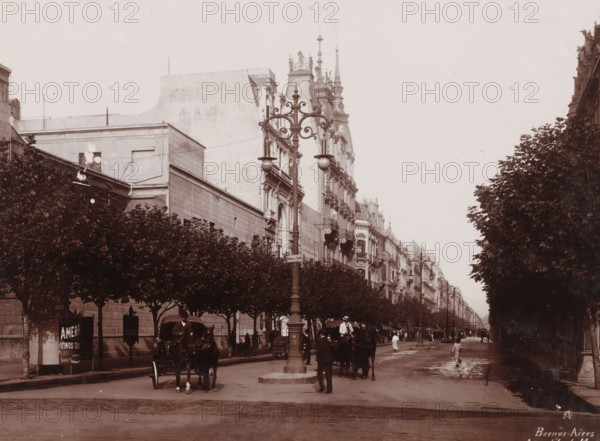 Buenos-Aires, Avenida de Mayo, View of Avenida de Mayo in Buenos Aires, photograph, monogram: 'AC', inscription: 'Buenos Aires 1904, 5', photographer: anonymous, Buenos Aires, 1904 - 1905, paper, pasteboard, baryta paper, daylight collodion silver print, height 166 mm x width 225 mm, height 259 mm x width 334 mm, city-view, and landscape with man-made constructions, A wide city street lined with tall, ornate buildings on one side and rows of trimmed trees on both sides; a decorative lamppost with curved metalwork stands near the center foreground, flanked by horse-drawn carriages and people walking or standing on the wet road; the facades of multi-story buildings show balconies, sculpted details, and steep rooflines with spires, while the tree canopies form a dark, leafy border along the avenue and cast shadows onto the sidewalk.