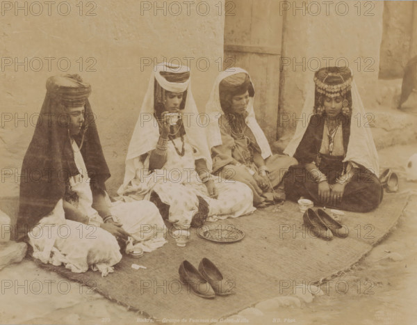 Biskra, groupe de femmes des Ouled-Naïls, Group portrait of four unknown Ouled Naïl women in Algeria, photograph, number: '299', photographer: Neurdein Frères, Algeria, 1863 - 1900, North Africa, pasteboard, paper, albumen print, height 278 mm x width 345 mm, anonymous historical persons depicted in a group, in a group-portrait - BB - woman, Four seated women wear layered garments and head coverings, with two wearing veils that drape over their shoulders and one wearing a decorative headpiece with hanging ornaments; they sit on a woven mat against a plastered wall and a wooden door, with various cups and a patterned tray placed on the mat in front of them and pairs of shoes positioned nearby.