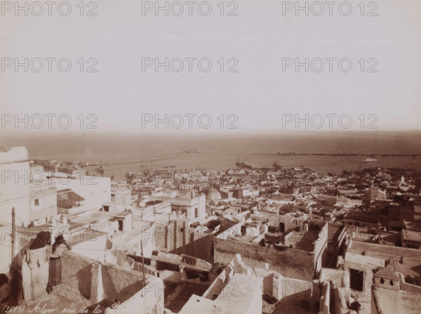 Alger, pris de la Casbah, View of Algiers and the Mediterranean from the Kasbah of Algiers, photograph, number: '(2609).', Kasbah of Algiers, 1860 - 1900, photographer, North Africa, Algiers, paper, albumen print, albumendruk, height 183 mm x width 247 mm, city-view in general, 'veduta', Mediterranean Sea, A wide panoramic view showing a dense cluster of flat-roofed buildings and rooftops in light tones descending toward a harbor, with laundry and cloths hanging on lines across several terraces, two figures leaning on a low parapet in the lower left, narrow alleys and stairways between the buildings, domed and rectangular roof structures scattered among the blocks, a calm sea stretching across the background with piers and several boats and ships moored or anchored near long breakwaters, and a pale sky occupying the upper portion of the scene.