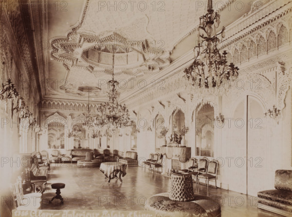 Alger, interior Palais d'hiver, Interior of the Dar Hassan Pacha in the Kasbah of Algiers, photograph, number: '2386', Kasbah of Algiers, 1860 - 1900, photographer, North Africa, Algiers, paper, albumen print, height 189 mm x width 250 mm, palace, Dar Hassan Pacha, A long ornate room with a high carved ceiling and multiple decorative chandeliers suspended along its length, walls covered with intricate carved arches and ornamental moldings, rows of upholstered chairs and sofas arranged along the sides, several round cushioned ottomans and small tables draped with cloths placed on a polished floor, wall sconces with hanging ornaments, a console table with decorative vases and objects beneath a mirror, and a large carved door set into one wall.