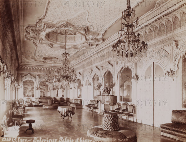 Interior of the Dar Hassan Pacha in the Kasbah of Algiers, Alger, interior Palais d'hiver, photograph, number: '2386', Kasbah of Algiers, 1860 - 1900, photographer, North Africa, Algiers, paper, albumen print, height 188 mm x width 246 mm, palace, Dar Hassan Pacha, A long ornate interior room with a highly detailed carved ceiling and decorative cornices, large crystal chandeliers hanging from the ceiling, tall arched mirrors and archways with intricate carved patterns along the walls, rows of upholstered chairs and settees arranged along the sides, round cushioned ottomans and small tables with cloth coverings on the polished floor, decorative vases and a sculptural centerpiece on a covered console, wall-mounted candle sconces, and richly patterned upholstery and fabrics throughout the furnishings.