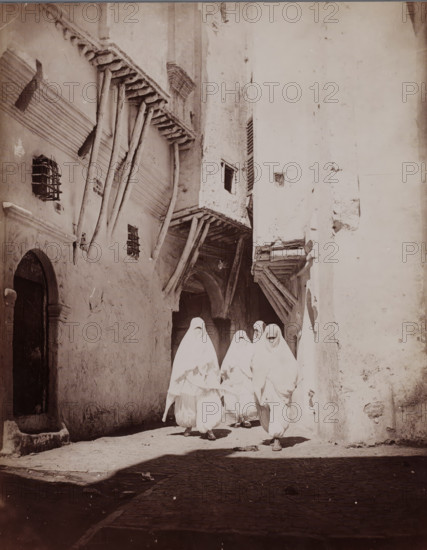 Alger (Rue Arabe), Four unknown veiled women on the street in Algiers, photograph, number: '2018.', Algiers, 1860 - 1900, photographer, North Africa, paper, albumen print, height 244 mm x width 187 mm, adult woman, street, A narrow sunlit alley flanked by tall, weathered buildings with rough plaster walls and projecting wooden beams, showing small barred windows and an arched doorway on the left, and timbered overhangs and recessed openings above; in the foreground a group of figures draped in light-colored garments covering their heads and bodies walk toward the camera, their faces mostly obscured, casting shadows on the cobbled ground.