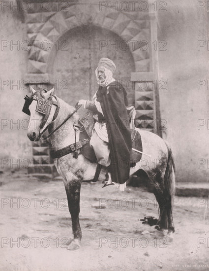Officer on horseback, probably in Algiers, Algeria, photomechanical print, inscription: 'Officier A...', photographer: Alexandre Leroux, Algiers, cliché maker: anonymous, 1880 - 1900, North Africa, paper, collotype, photoprint, height 265 mm x width 209 mm, horse, officer, A person wearing layered garments and a wrapped head covering sits astride a horse outfitted with ornate tack and decorative coverings on its head and body, positioned in front of a stone wall with a large arched recess and patterned masonry; the ground beneath them appears uneven and textured, and the scene is rendered in a monochrome palette.
