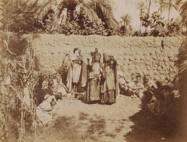 Danse des Ouled Naïls, Group Ouled Naïl people in Algeria, Types Algeriens, photograph, number: 'L. 259', photographer: Neurdein Frères, Algeria, c. 1863 - 1900, North Africa, pasteboard, paper, albumen print, height 278 mm x width 345 mm, anonymous historical persons depicted in a group, in a group-portrait, A group of people arranged outdoors in front of a textured earthen wall with tall palm fronds and foliage visible above it, with two women standing near the center wearing ornate headdresses and long layered garments while other figures sit or crouch around them on the ground, some wrapped in cloaks or head coverings, with a person standing slightly behind the central pair wearing a draped robe and headwrap and other seated individuals forming a loose semicircle, dried plants and stalks along the left edge, scattered pottery or small vessels on the ground, and patches of sunlit bare earth and grassy tufts in the foreground.