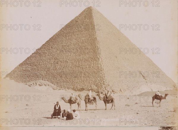 View of the Pyramids of Giza, photograph, Giza, 1860 - 1900, photographer, North Africa, pasteboard, paper, albumen print, height 190 mm x width 278 mm, height 396 mm x width 290 mm, pyramid (historical grave form), Pyramids of Giza, A large stone pyramid fills the background with its stepped blocks visible on two faces, and a sandy foreground shows a group of people and camels; some riders sit atop camels wearing loose garments and head coverings while others sit on the ground in a small cluster, and one person sits alone facing the camera.