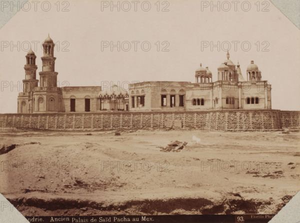Alexandrie, ancien palais de Saïd Pacha au Mex, View of the palace of Mohammed Said at the port of Alexandria, photograph, number: '93.', photographer: Luigi Fiorillo, Alexandria, 1870 - 1890, North Africa, pasteboard, paper, albumen print, height 316 mm x width 404 mm, Person, Mohammed Said (governor of Egypt), palace, Palace of Mohammed Said, A wide, sepia-toned photograph shows a large stone building complex behind a textured stone wall, with multiple domes and ornate towers rising above the roofline; two tall cylindrical towers with open-arched upper sections stand on the left, while a variety of rounded domes and small cupola-like structures appear toward the right, some supported by slender columns. The façade of the buildings features rectangular and arched openings, some appearing dark and empty, and decorative circular and arched window frames on portions of the walls. In front of the wall is a barren, uneven expanse of ground with scattered low vegetation and shallow depressi...