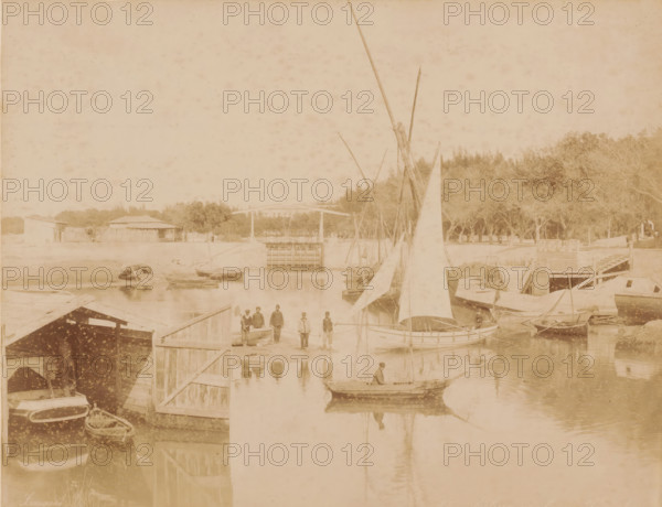 Sailboats at the lock of Ismailia, Ecluse arab a Ismailia, photograph, inscription: 'foto Zangaki, Arab lock in Ismailia', photographer: Zangaki, C. & G., Ismailia, 1870 - 1910, North Africa, pasteboard, paper, albumen print, height 216 mm x width 279 mm, height 348 mm x width 454 mm, sailing-ship, sailing-boat, sluice, weir, A waterfront scene with several small sailboats and rowboats moored along wooden docks and piers, one boat in the foreground with a person seated inside, a group of people standing on a dock near the center, tall masts and sails rising from boats clustered along the water, reflections of boats and people visible on the water surface, wooden boathouses and platforms lining the shore, a tree-lined embankment behind the docks, and a low building with a pitched roof visible beyond the trees.