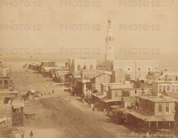 Port Saïd Village arabe, View of Port Said, photograph, inscription: 'photo Zangaki, Port Said Arab village', number: 'No. 451', photographer: Zangaki, C. & G., Port Said, 1870 - 1910, North Africa, pasteboard, paper, albumen print, height 220 mm x width 278 mm, height 348 mm x width 454 mm, public road in village, temple, shrine, Islam, Mohammedanism, A sepia-toned street scene with rows of wooden and masonry buildings lining both sides of a wide unpaved road that leads toward a shoreline, a tall minaret rising above a large striped building near the water, small awnings and balconies projecting from many structures, scattered groups of people walking or standing along the street, carts and market stalls visible near the foreground, and the calm sea forming a horizontal band on the horizon.