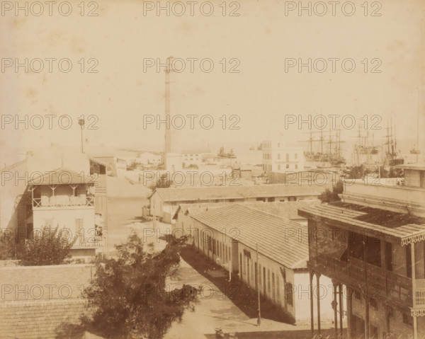 Rade de Port Said, View of the harbor of Port Said, Egypt, photograph, inscription: 'foto Rives, rade Port Said', inscription: 'BKF Rives', number: 'No. 15 ?', 1851 - 1900, photographer, North Africa, pasteboard, paper, albumen print, height 349 mm x width 454 mm, harbour, Port Said, A sepia-toned coastal scene showing a row of low buildings with tiled roofs extending toward a waterfront, a tall slender lighthouse rising near the center with a small structure at its top, a multi-story building near the water with rectangular windows and a flagpole, several masted sailing ships moored in the harbor to the right, wooden balconies and verandas on foreground buildings, a narrow street running between the structures with scattered trees and shadows, and a calm expanse of sea visible beyond the waterfront.