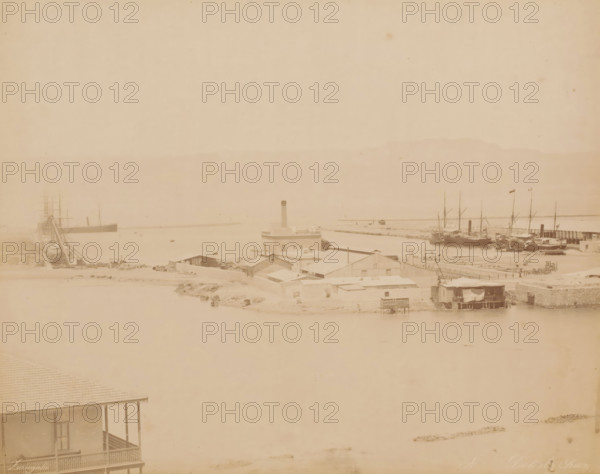 Dok de Suez, Dock at the Suez Canal, photograph, number: 'No', inscription: 'foto Zangaki, Suez', photographer: Zangaki, C. & G., Suez Canal, 1870 - 1910, North Africa, pasteboard, paper, albumen print, height 349 mm x width 454 mm, canal, dry-dock, floating-dock, A faded panoramic view of a harbor area showing a stretch of calm water bordered by low buildings and docks, a cluster of ships moored at the right with tall masts and flags, another large vessel at the left with visible rigging, a central blocky building with a tall chimney rising above surrounding single-story structures, assorted small sheds and waterfront platforms, and a foreground porch with a decorative railing partially visible at the lower left.