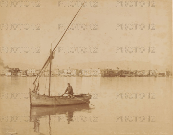 Panorama de Suez et de ..., Sailboat on the Suez Canal, photograph, inscription: 'foto Zangaki, Panorama Suez', number: 'No 17', photographer: Zangaki, C. & G., Suez Canal, 1870 - 1910, North Africa, pasteboard, paper, albumen print, height 349 mm x width 454 mm, sailing-ship, sailing-boat, canal, A sepia-toned scene showing a small wooden sailboat floating on calm water with a single standing man holding an oar near the center of the boat, a tall mast with furled sails and rigging rising upward, and the boat's reflection visible on the water; in the distant background a low waterfront with a row of buildings and assorted small boats along the shore stretches across the image under a pale sky.