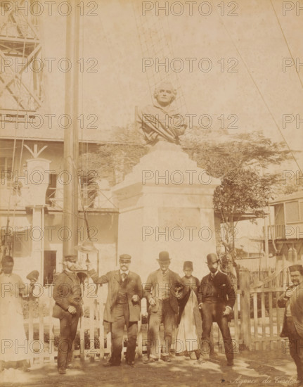 Six men and a woman standing in front of a monument, probably in Egypt, Agence du Terelain, photograph, inscription: 'foto Zangaki', photographer: Zangaki, C. & G., Egypt, 1870 - 1910, North Africa, pasteboard, paper, albumen print, height 453 mm x width 348 mm, monument, statue, A large stone bust on a stepped pedestal dominates the center of the scene, with a tall pole and rigging rising behind it and a netted structure visible in the background; a row of men and a woman stand in front of the pedestal along a low picket fence, several of the men wearing formal coats and hats and some holding their hats in hand, while the woman wears a long dress and head covering; buildings with balconies, windows, and wooden railings form the background on either side, and small trees and shrubbery are visible near the pedestal and along the fence.