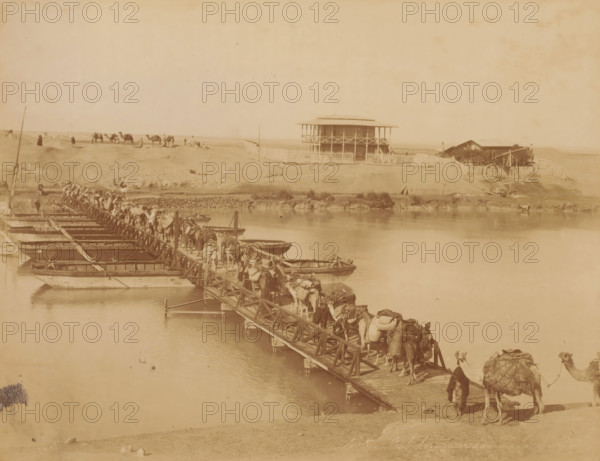 Caravan with camels or dromedaries on a pontoon bridge, Pont des caravannes de la ..., photograph, number: 'No ...', photographer: Zangaki, C. & G., 1870 - 1910, pasteboard, paper, albumen print, albumendruk, height 348 mm x width 454 mm, hoofed animals: camel, pontoon bridge, A sepia-toned riverside scene shows a long wooden pontoon bridge extending across calm water, with numerous camels and people walking along it; small flatboats are moored alongside the bridge, and cargo is visible on some camels. On the far bank a two-story wooden building with a wraparound balcony and a fenced yard stands atop a low rise, with a smaller structure and vegetation nearby. In the background a sandy landscape stretches with scattered figures and a group of camels standing on the plain. The sky appears clear and pale above the horizon.
